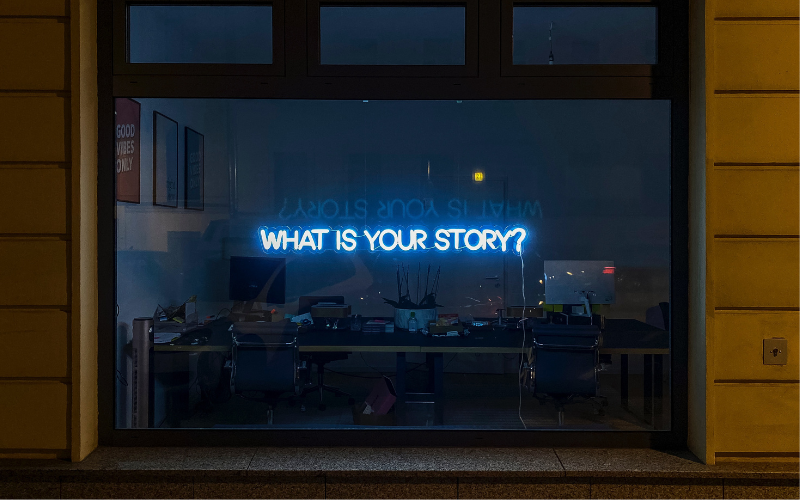 Neon sign in a window that says What Is Your Story?